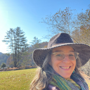 Woman in hat smiling outdoors with trees and clear blue sky in the background.