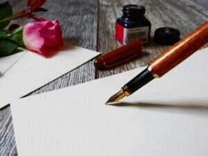 A fountain pen on paper with a rose, an ink bottle, and cap on a wooden surface.