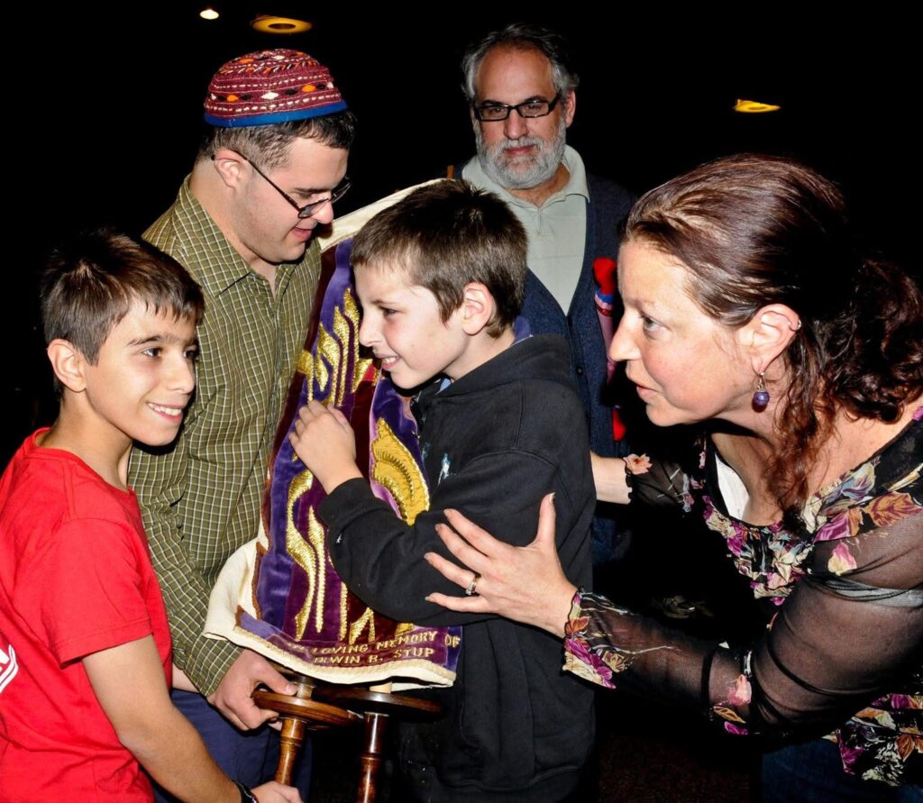 People gather around a boy holding a Torah scroll, engaged in a joyful interaction.