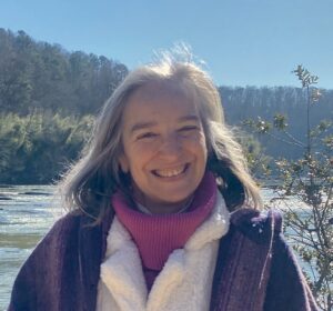 McKenzie is a woman with long gray hair smiles, standing outdoors near a river with trees in the background.