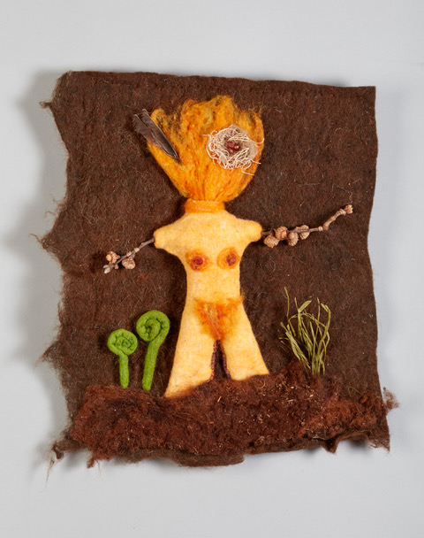 Textile art of a female figure with fiery hair, twig arms, and plants on a brown fabric background.