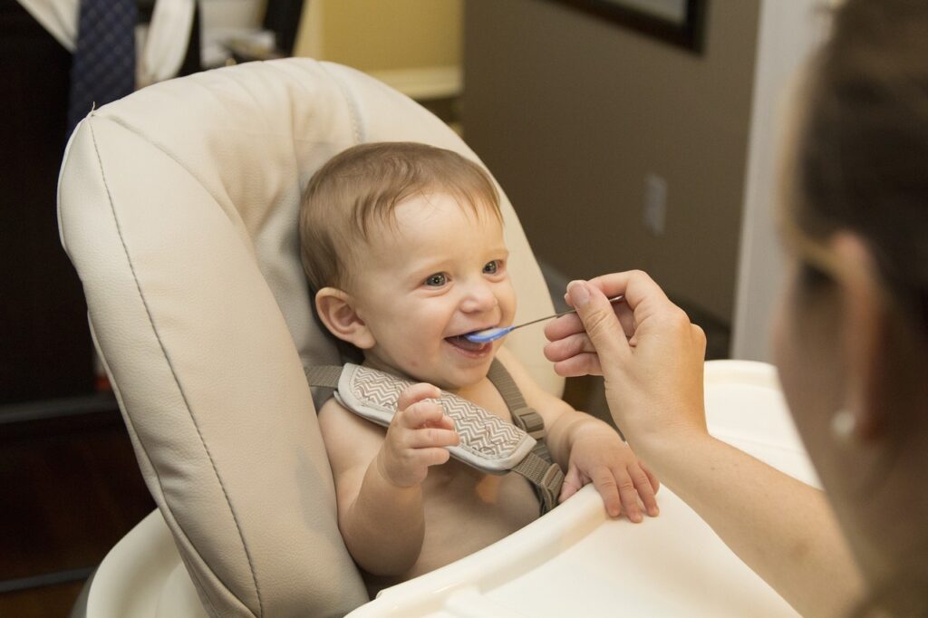 A smiling baby in a high chair being fed with a spoon by an adult.