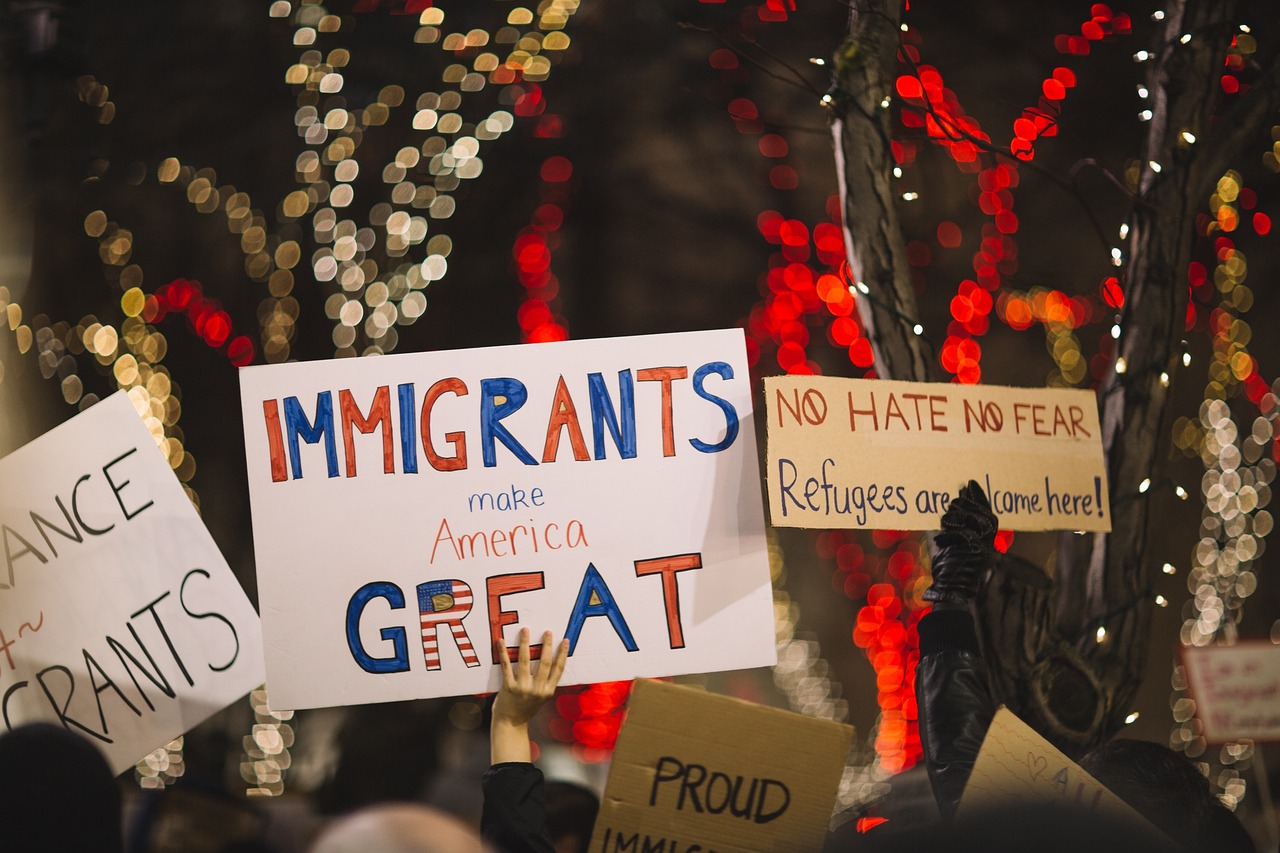 People holding signs supporting immigrants and refugees at a protest with festive lights in the background.