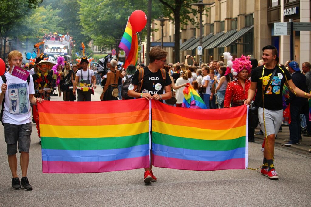 People holding a large rainbow flag during a vibrant outdoor pride parade with a colorful crowd in the background.