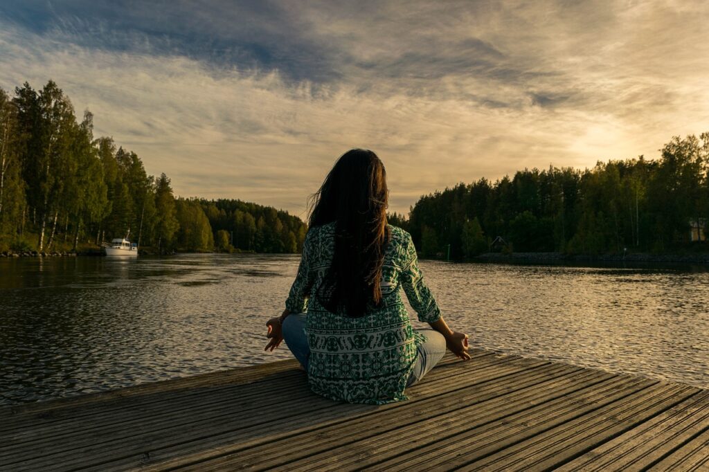 Person meditating on a wooden dock facing a lake and trees during sunset.