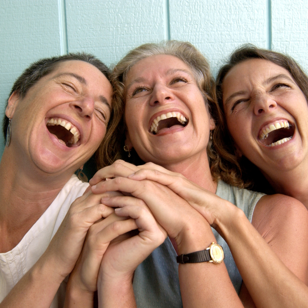 Three women laughing together, holding hands, against a light blue wall background.