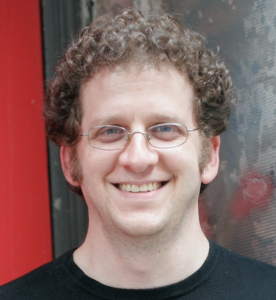 A person with curly hair, wearing glasses and a black shirt, smiles in front of a red and textured background.