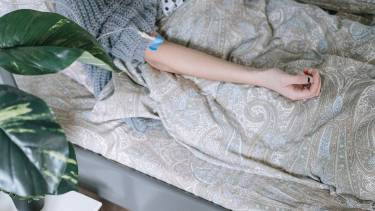 A person lying in bed with an IV in their arm, covered by a patterned blanket, next to a leafy green plant.