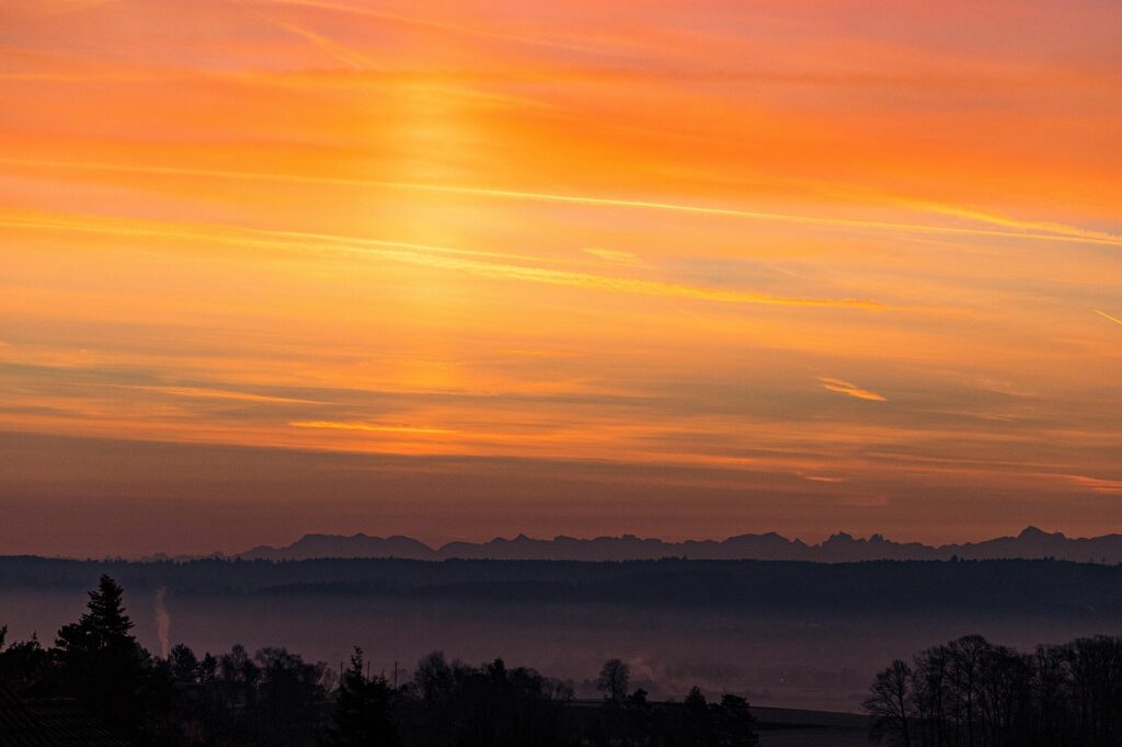 Orange and pink sunset over distant mountain silhouette with a misty foreground of trees.