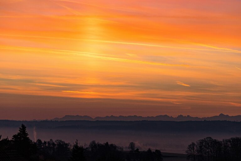 Orange and pink sunset over distant mountain silhouette with a misty foreground of trees.