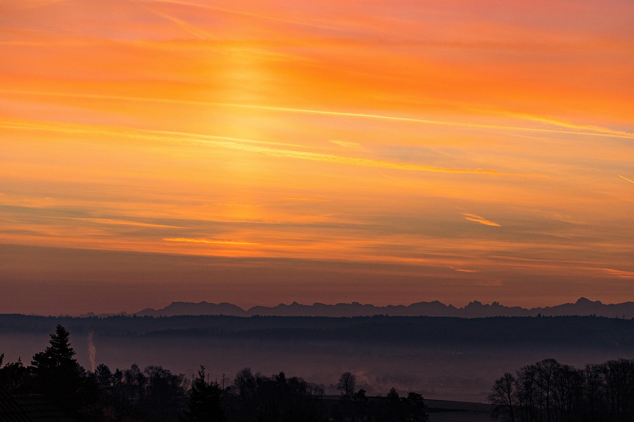 Orange and pink sunset over distant mountain silhouette with a misty foreground of trees.