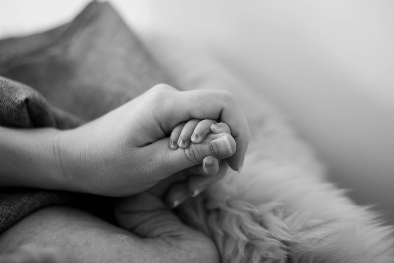 Close-up of an adult hand gently holding a babys hand, both resting on a soft, fluffy surface. Black and white.