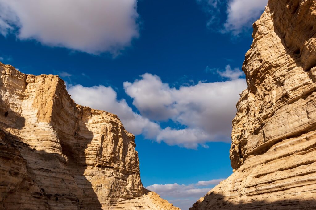 Desert canyon with towering cliffs under a bright blue sky and scattered white clouds.