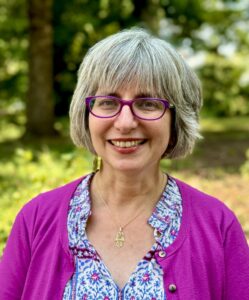 Smiling woman with gray hair, wearing purple glasses and a matching cardigan, outdoors with trees in the background.