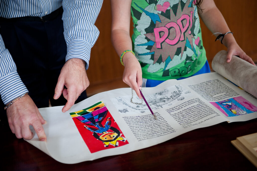 Two people point to illustrated and handwritten text on a scroll displayed on a table.