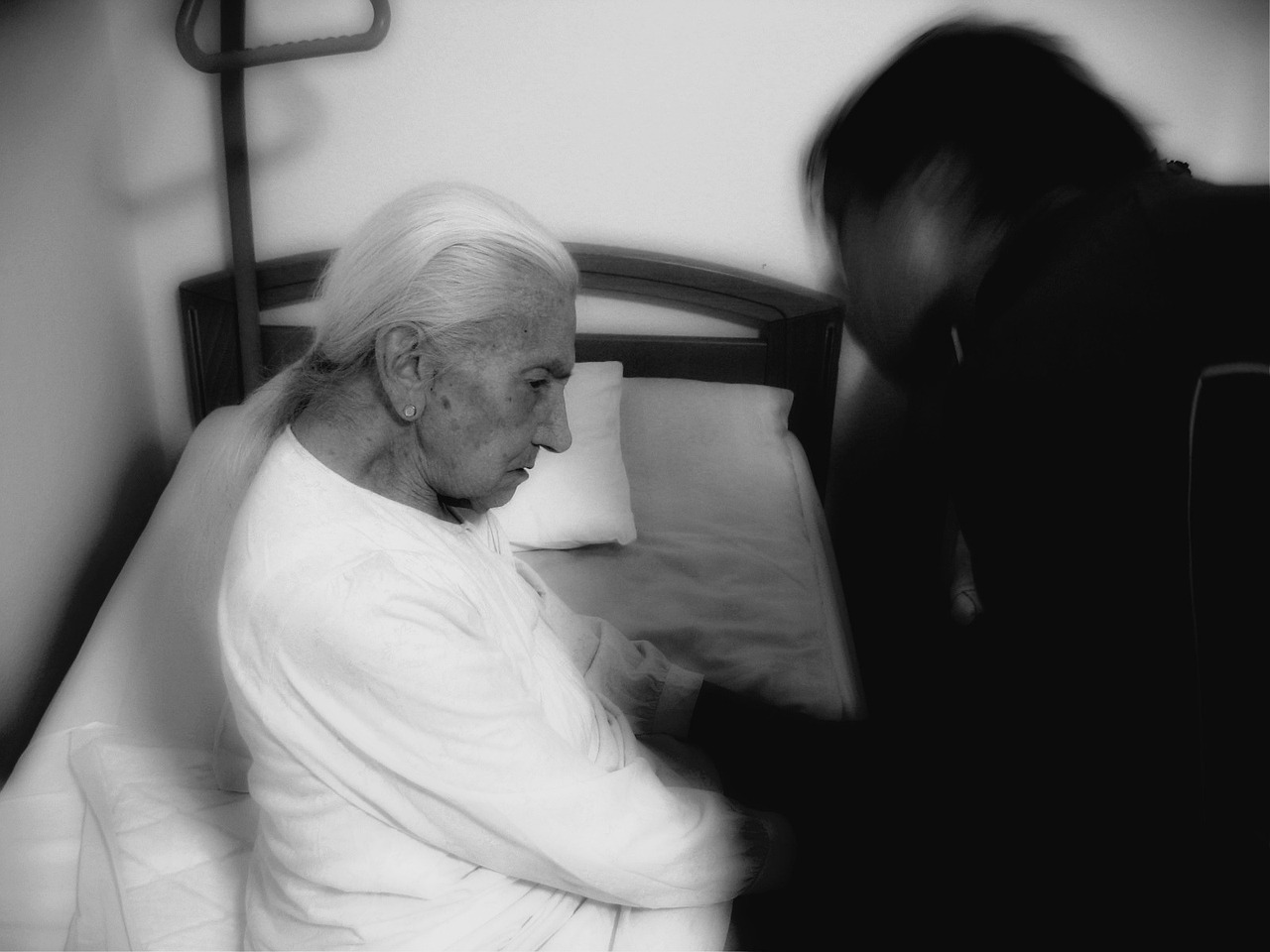 Elderly person sitting on a bed, with another person beside them in a dimly lit room.