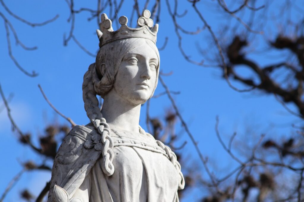 Stone statue of a crowned woman with braided hair against a blue sky and tree branches.