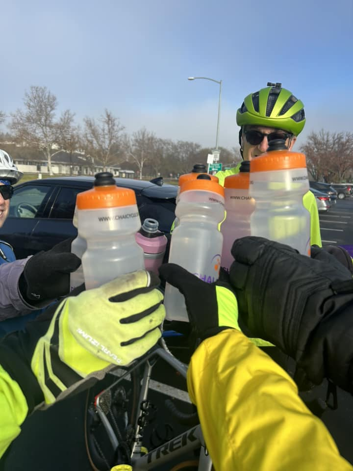 Cyclists wearing helmets toast with water bottles in a parking lot on a cloudy day.