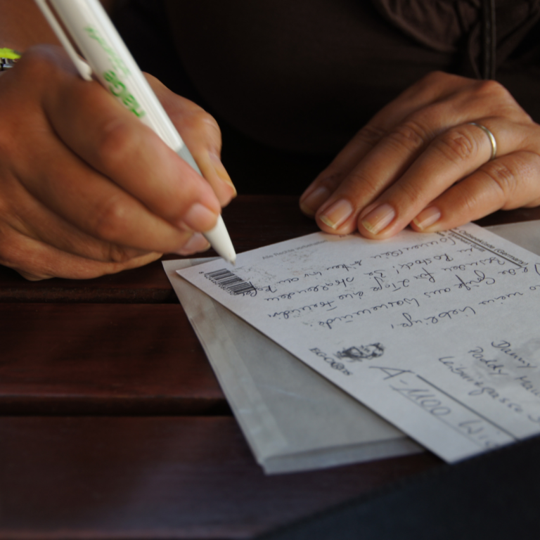 A person writes a message on a postcard with a pen at a wooden table.
