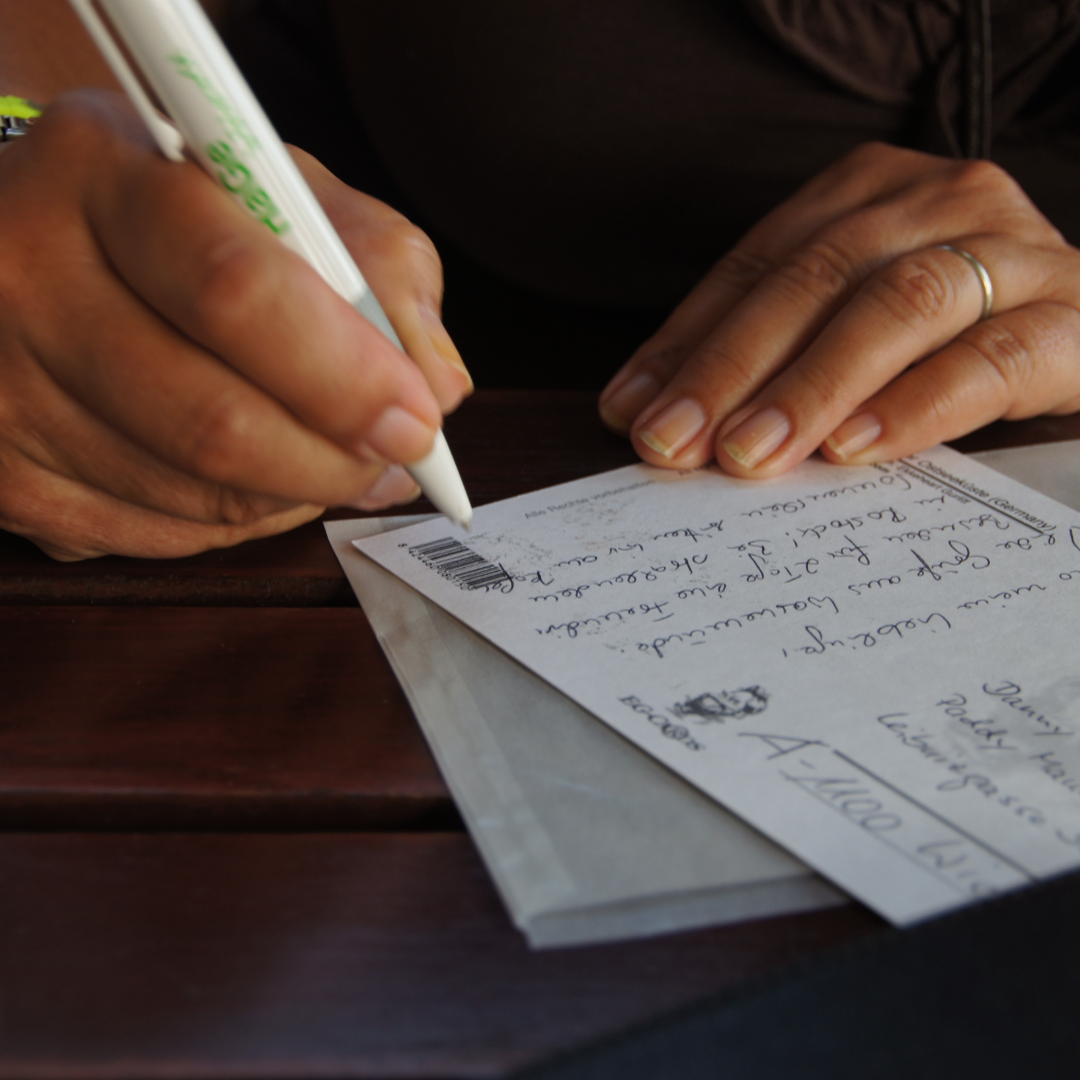 A person writes a message on a postcard with a pen at a wooden table.