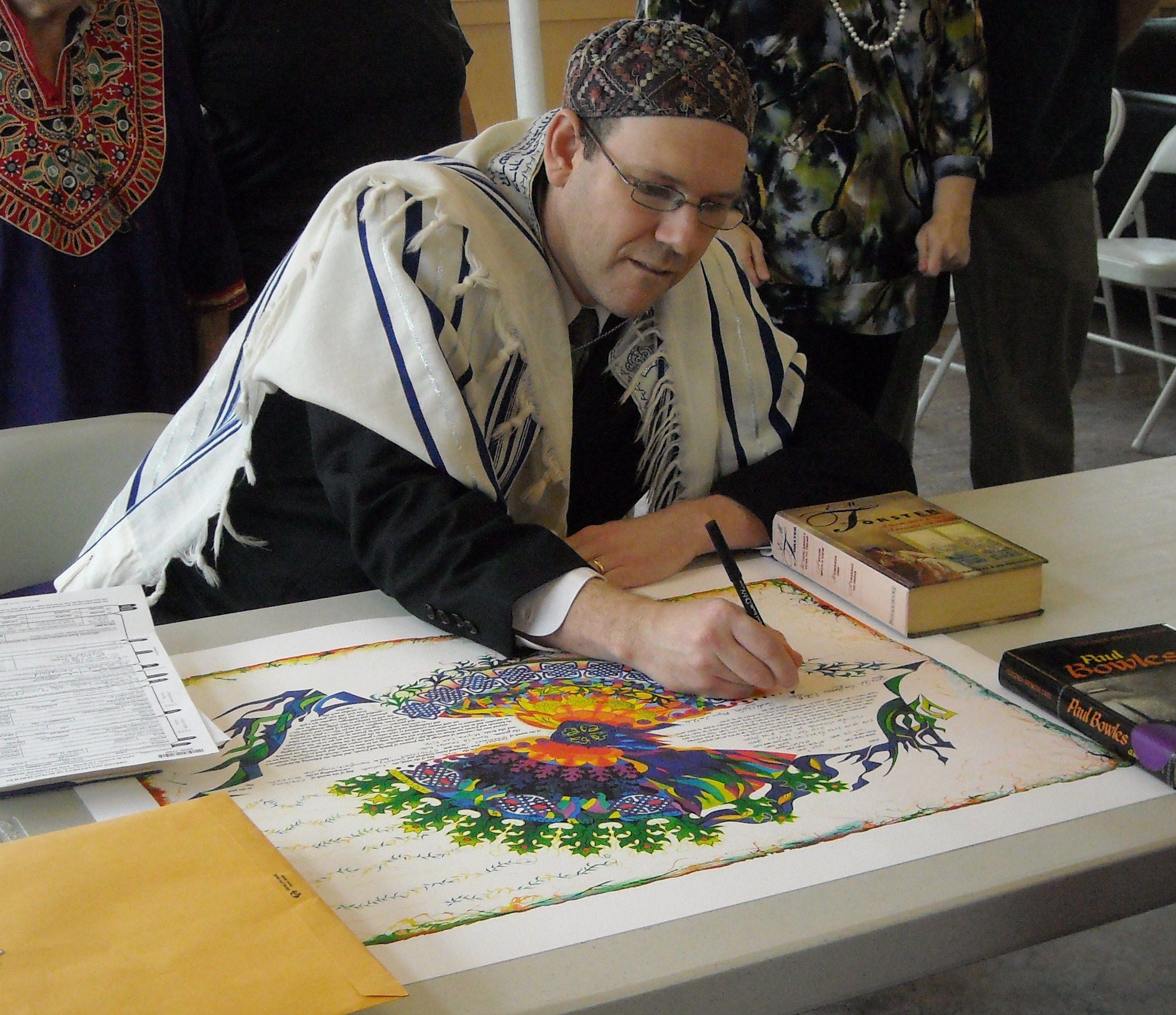 A man in religious attire signs a colorful document at a table with books and papers.