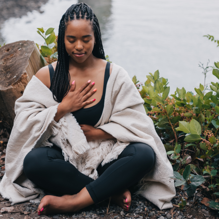 Woman sitting cross-legged by water, eyes closed, hand on chest, wrapped in a cozy blanket outdoors.