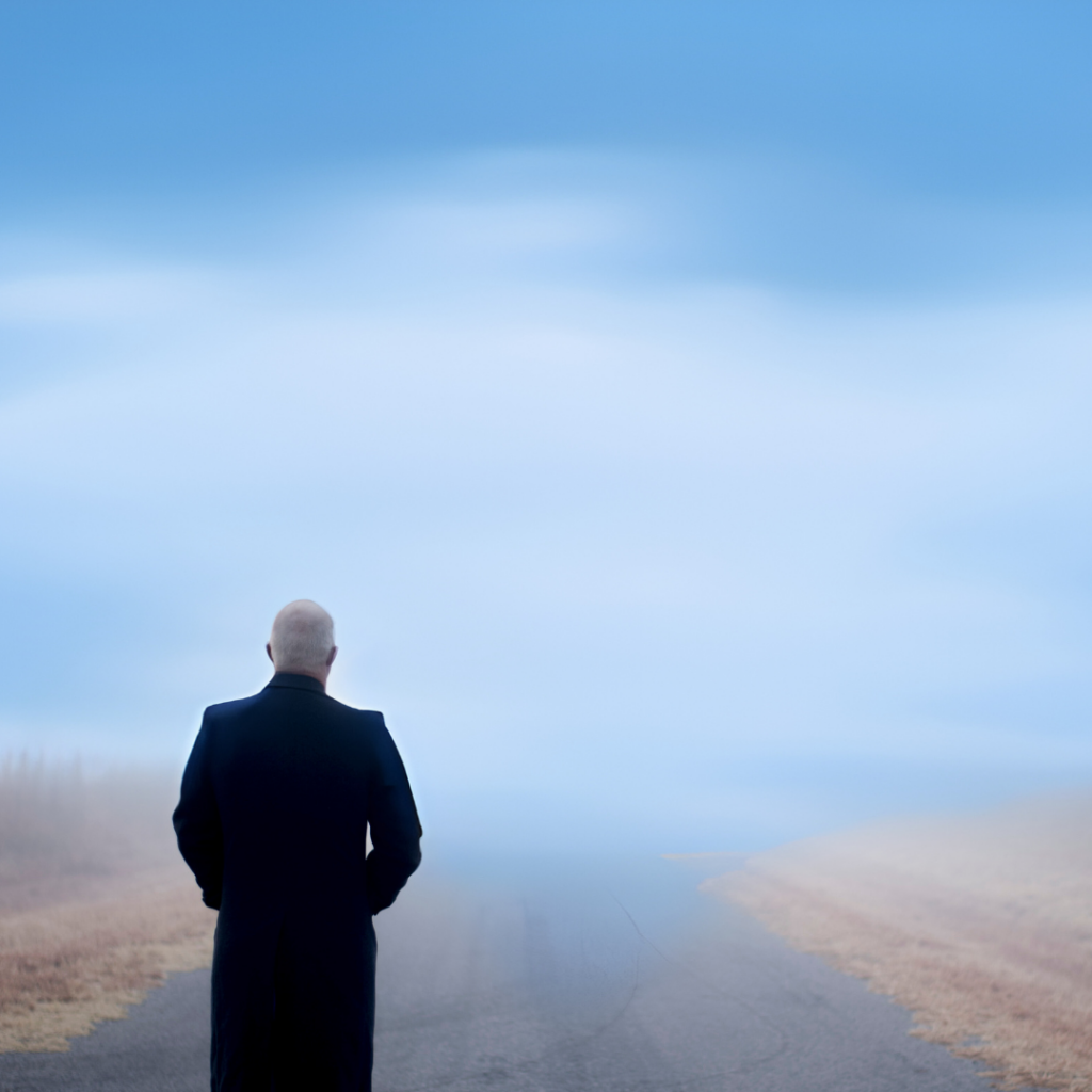 A person in a dark coat stands alone on a deserted road under a cloudy, blue sky.