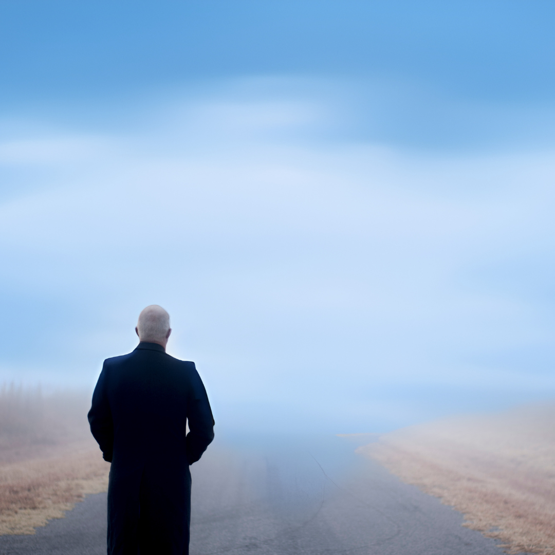 A person in a dark coat stands alone on a deserted road under a cloudy, blue sky.