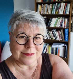 Smiling person with short gray hair and round glasses in front of a bookshelf filled with books.