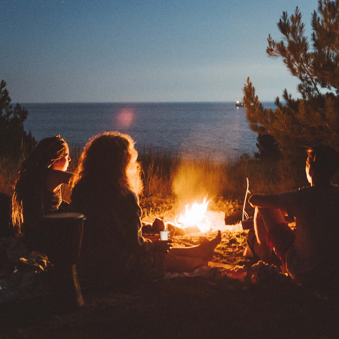 Three people sit around a campfire near the shore at dusk, with the sea and trees in the background.