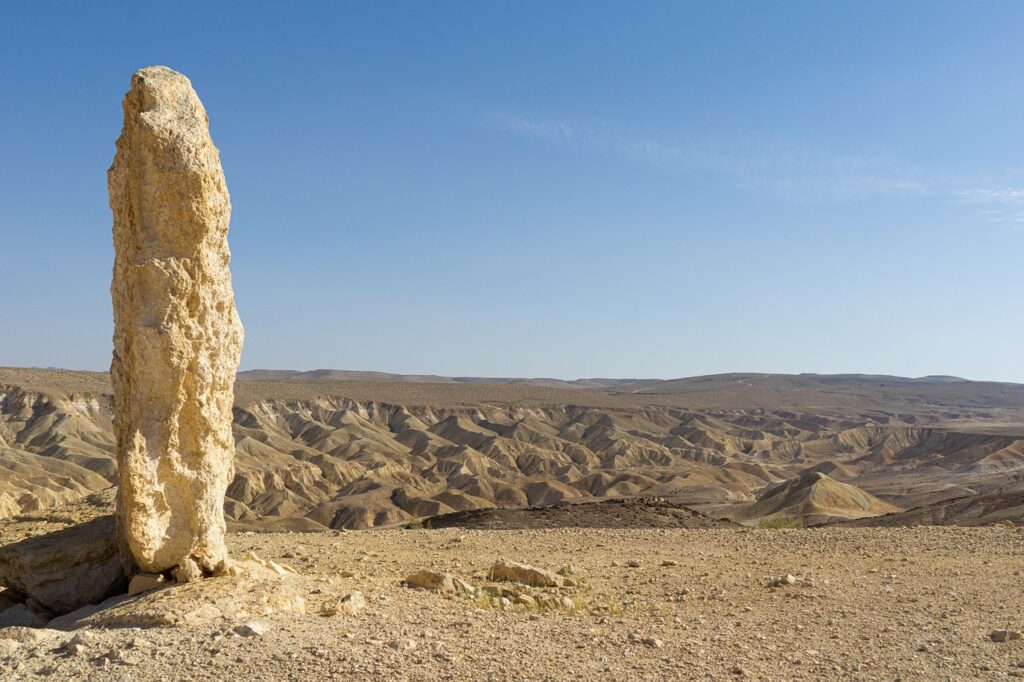 A tall, narrow rock stands in a dry, rocky desert landscape under a clear blue sky.