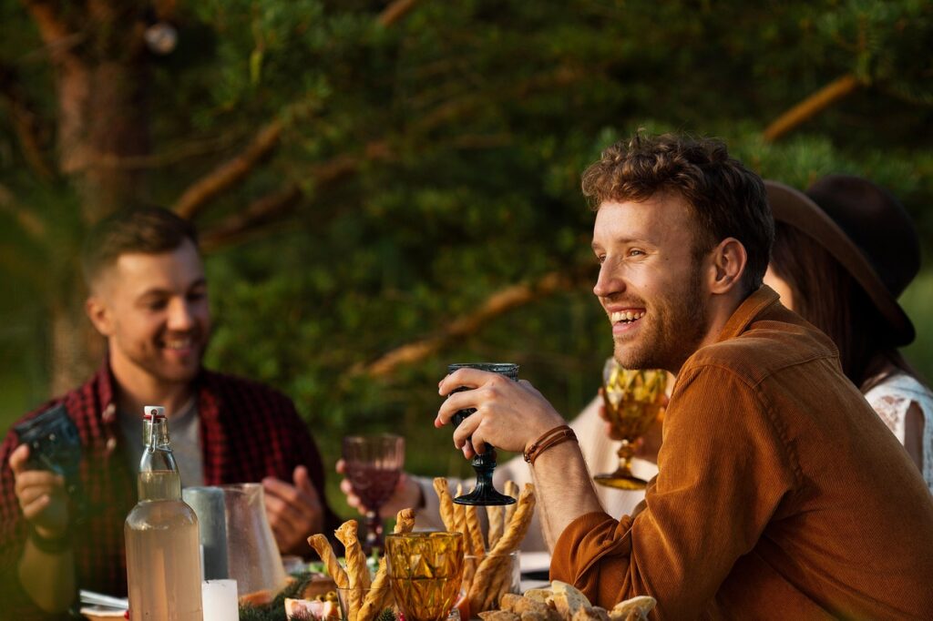 Three friends enjoying drinks and food outdoors at a table, smiling and laughing together in warm sunlight.