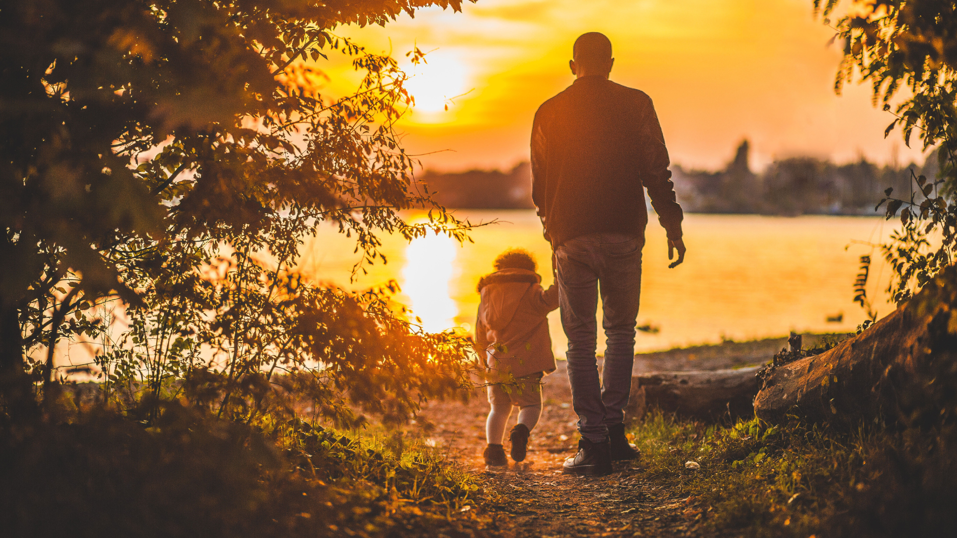 An adult and child walk by a lakeside at sunset, surrounded by trees and warm golden light.