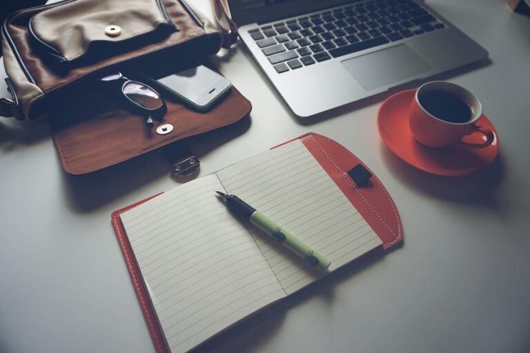 Open notebook with pen, laptop, coffee cup, glasses, and phone on a desk next to a leather bag.