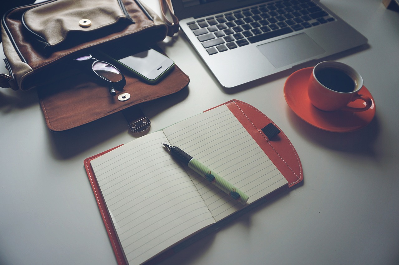 Open notebook with pen, laptop, coffee cup, glasses, and phone on a desk next to a leather bag.