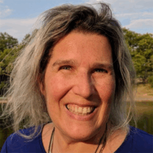Smiling woman with wavy gray hair outdoors, trees and water in the background.