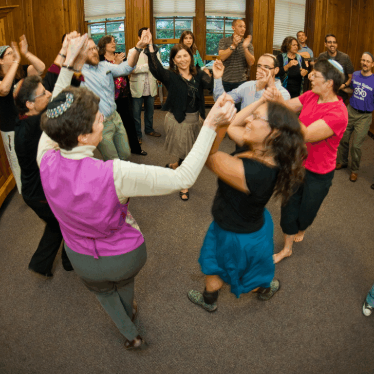 A group of people hold hands and dance in a lively circle inside a wood-paneled room.