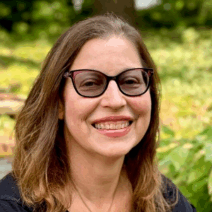 Smiling woman with long brown hair and glasses outdoors, with green foliage in the background.