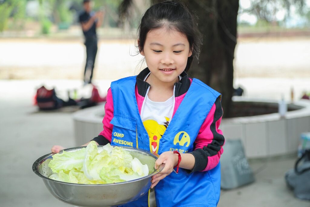 Young girl wearing a blue vest holds a large bowl of leafy lettuce outdoors, smiling.