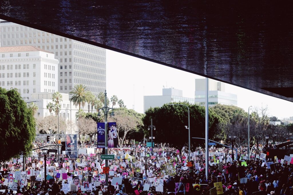A large crowd gathers outdoors holding colorful protest signs in a city surrounded by trees and tall buildings.