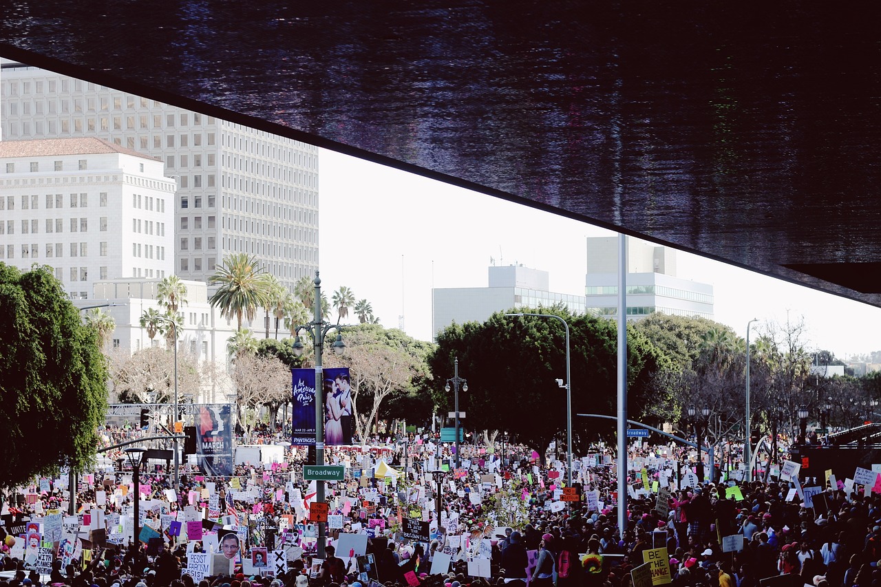 A large crowd gathers outdoors holding colorful protest signs in a city surrounded by trees and tall buildings.