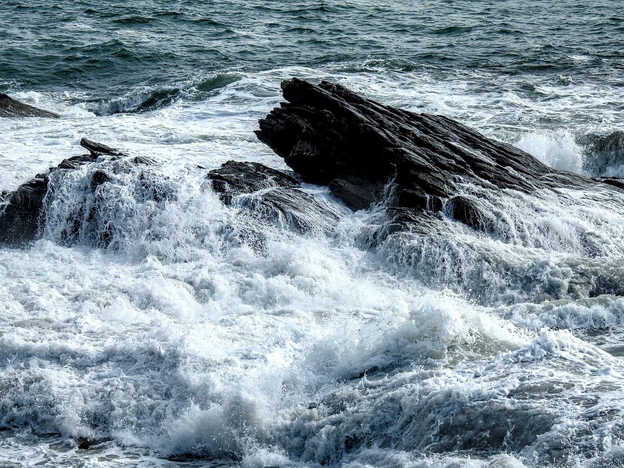 Waves crash against jagged dark rocks in the ocean, sending up white sea spray and foam.