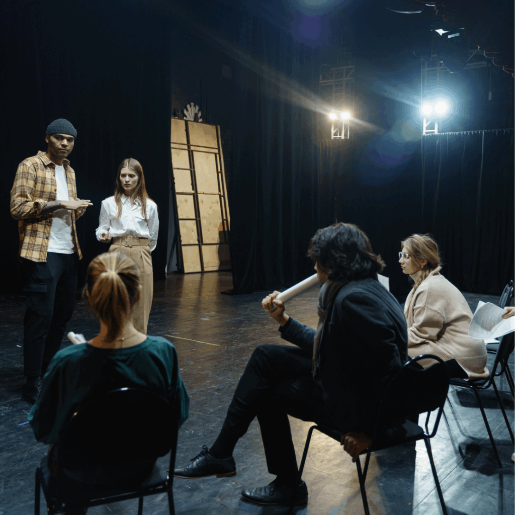 Two people stand on stage rehearsing while three others sit and watch, holding scripts in a dark theater.