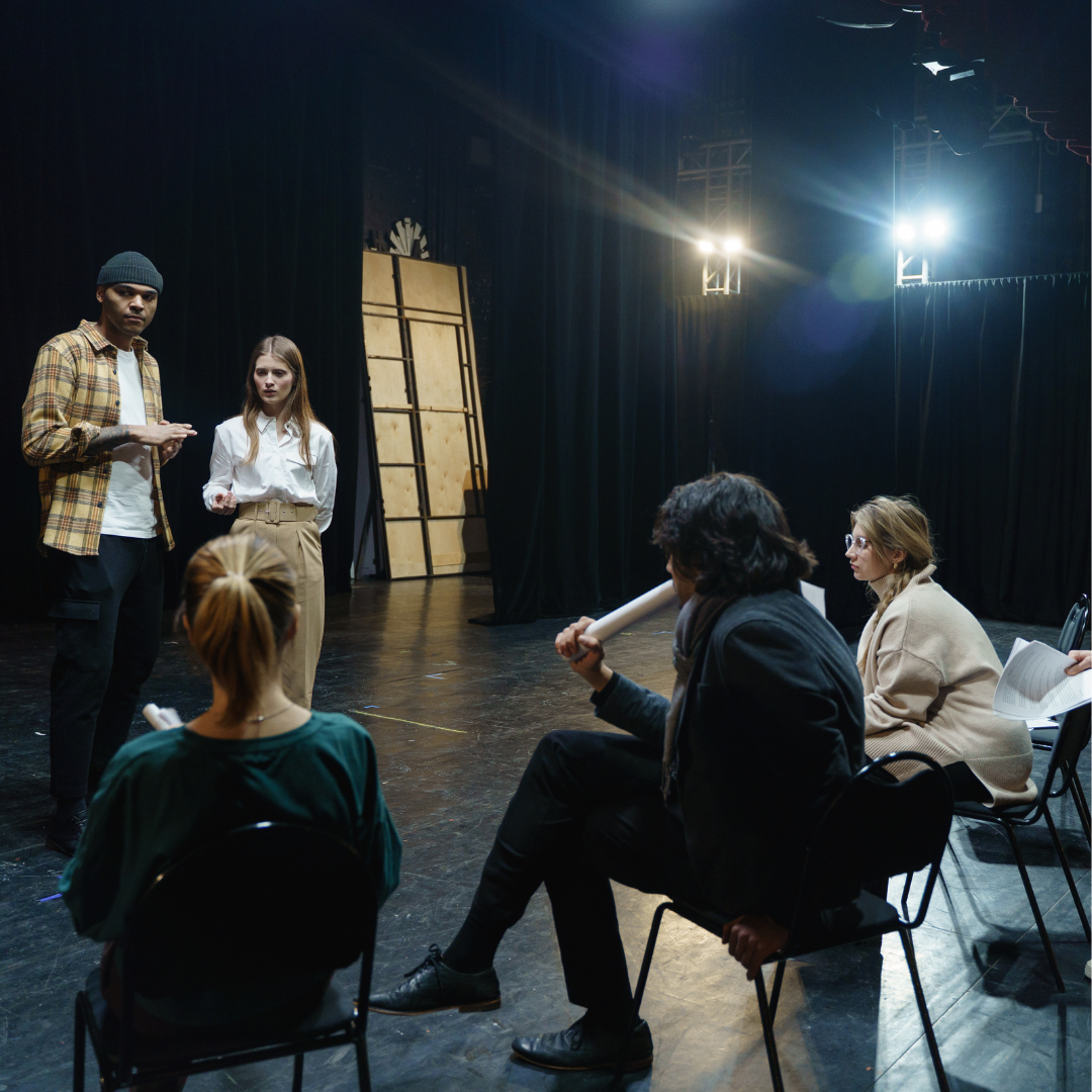 Two people stand on stage rehearsing while three others sit and watch, holding scripts in a dark theater.