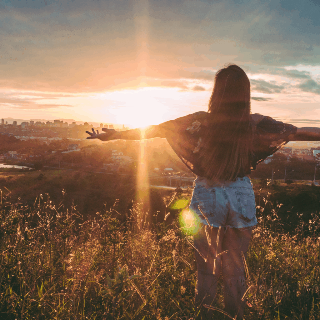 Woman standing in a field at sunset with arms outstretched, facing the sun and city skyline.