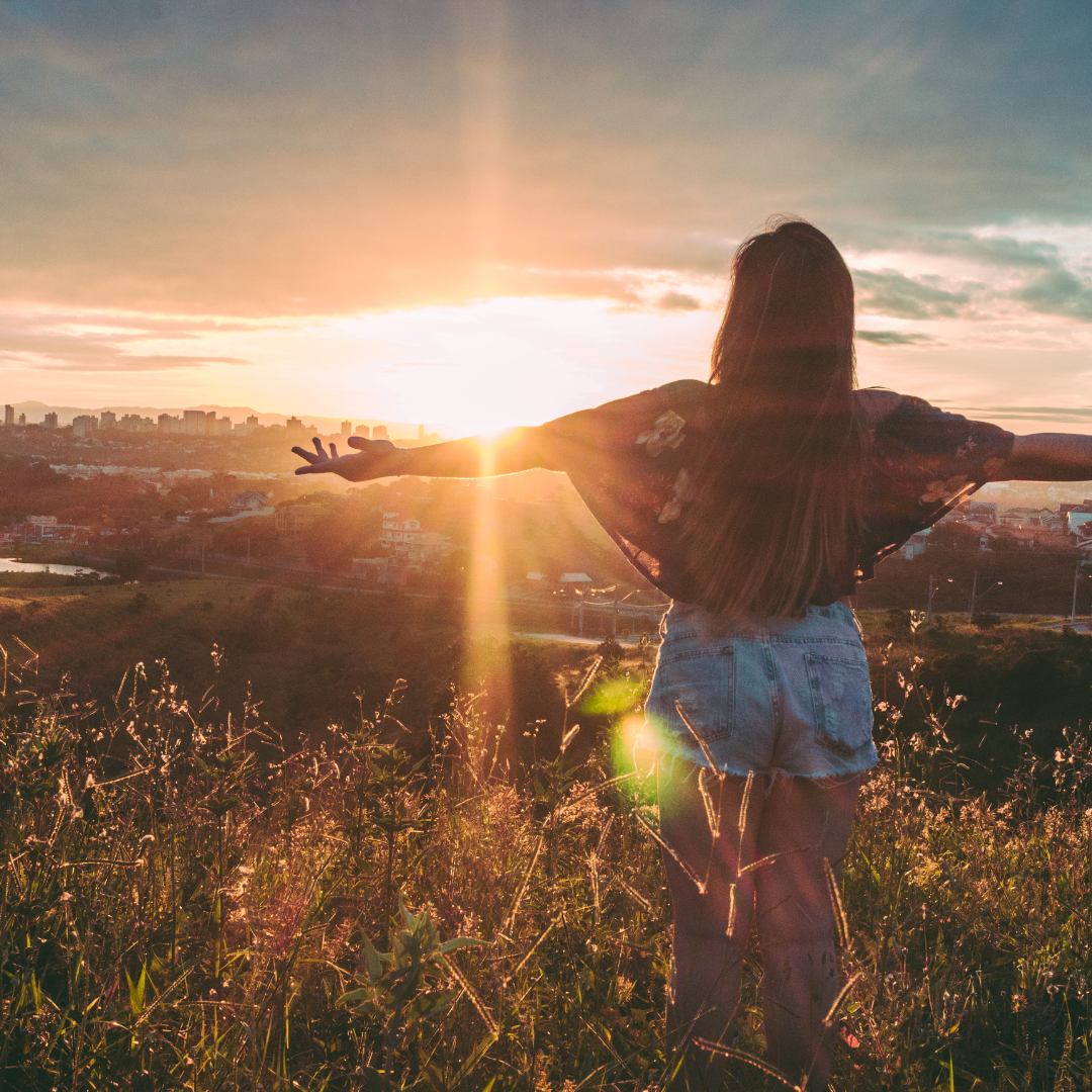 Woman standing in a field at sunset with arms outstretched, facing the sun and city skyline.