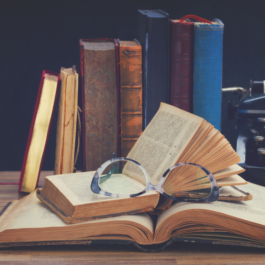 A pair of glasses rests on open books with old books stacked in the background on a wooden table.