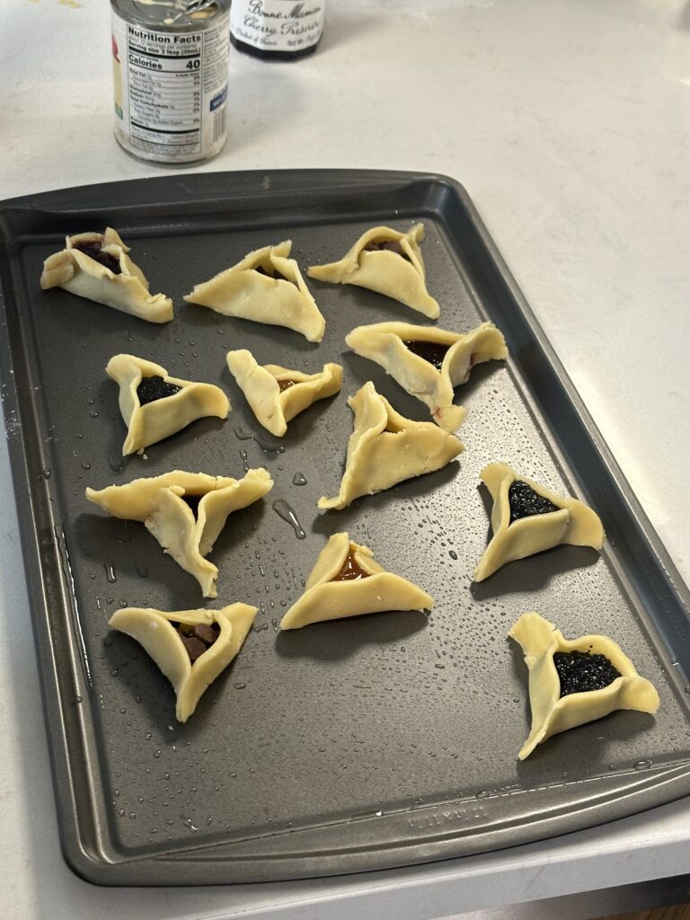Unbaked hamantaschen cookies with various fillings on a baking tray, ready to be baked.