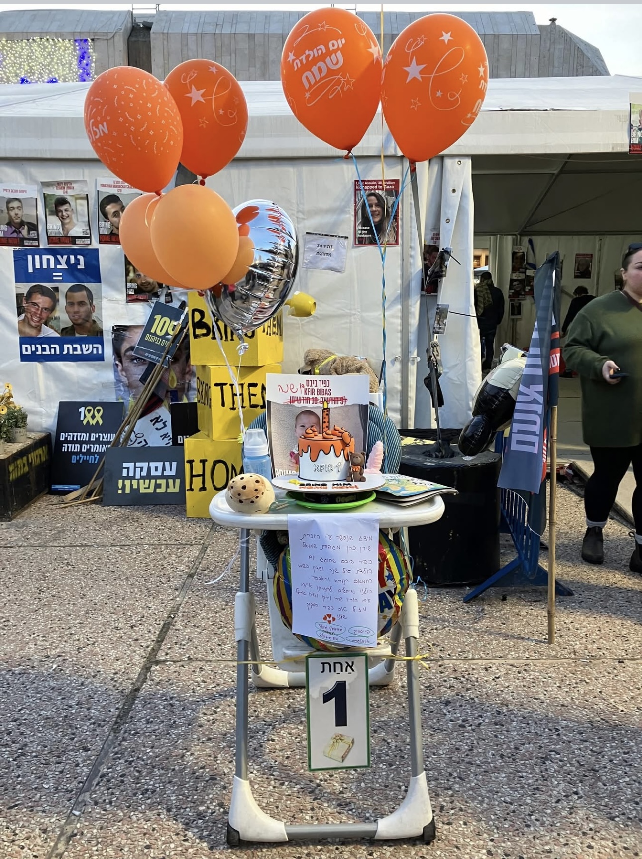 A highchair with orange balloons, protest signs, and a sign in Hebrew at an outdoor event with people nearby.