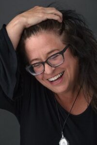 Woman with glasses smiling, wearing black top, hand in hair, dark background.